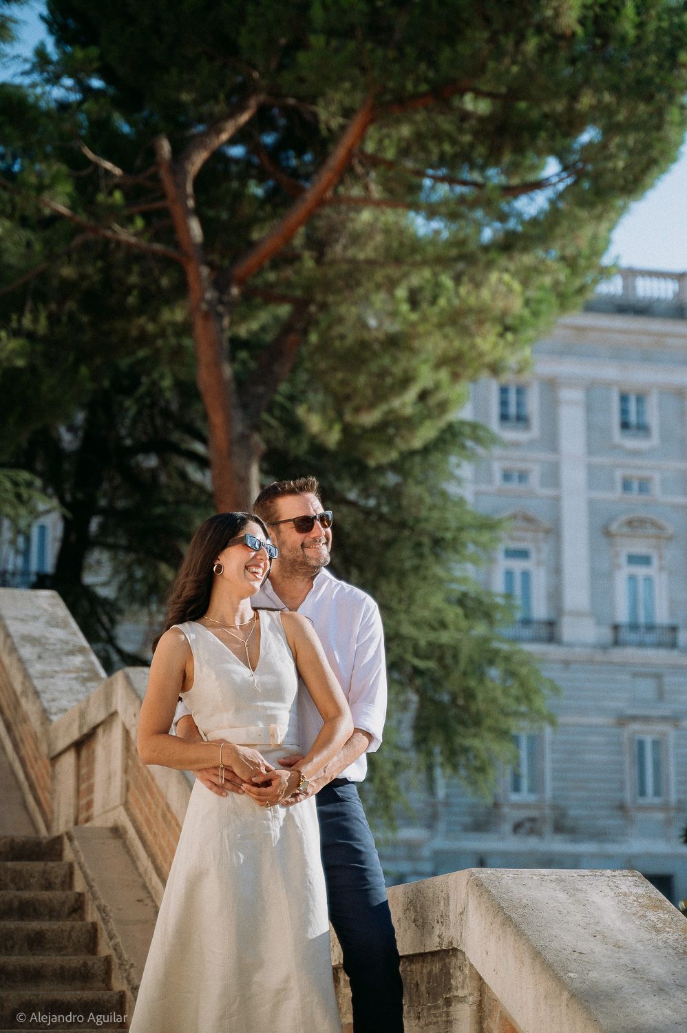 Couple in sunglasses embracing on stone steps near a grand building and pine tree in Madrid.