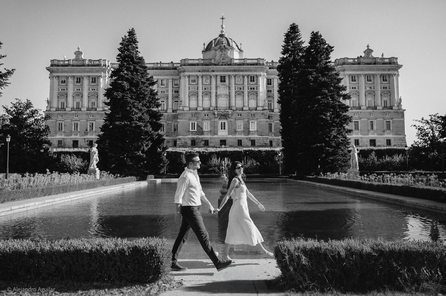 Black and white photo of a couple holding hands walking in front of the Royal Palace of Madrid.