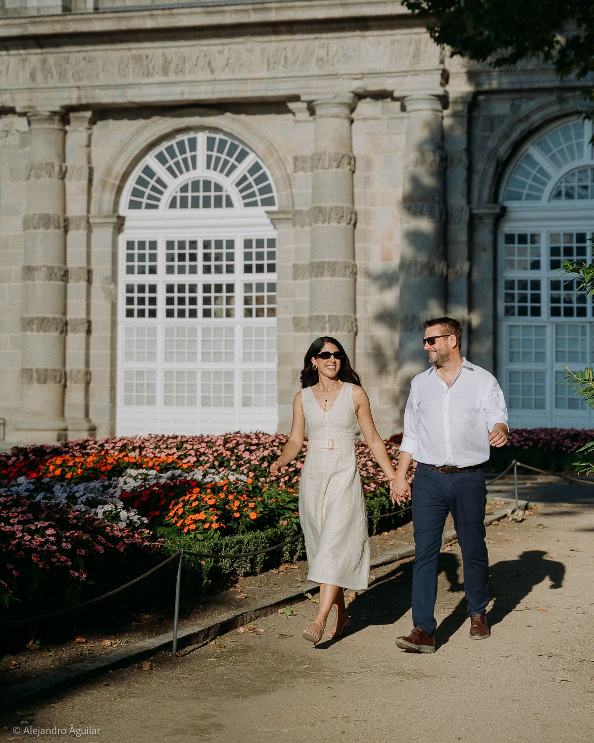 Couple holding hands walking past colorful flower beds outside a grand stone building on a sunny day.