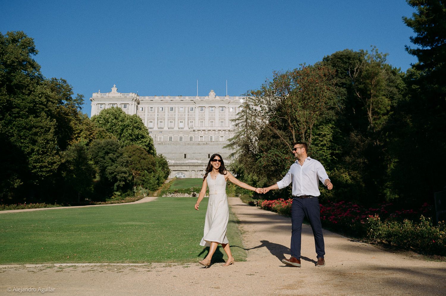 Couple holding hands in front of the Royal Palace of Madrid on a sunny day in lush green gardens.