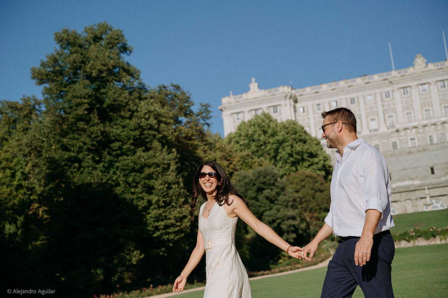 Couple holding hands walking on green lawn with historic palace and trees in background on sunny day.