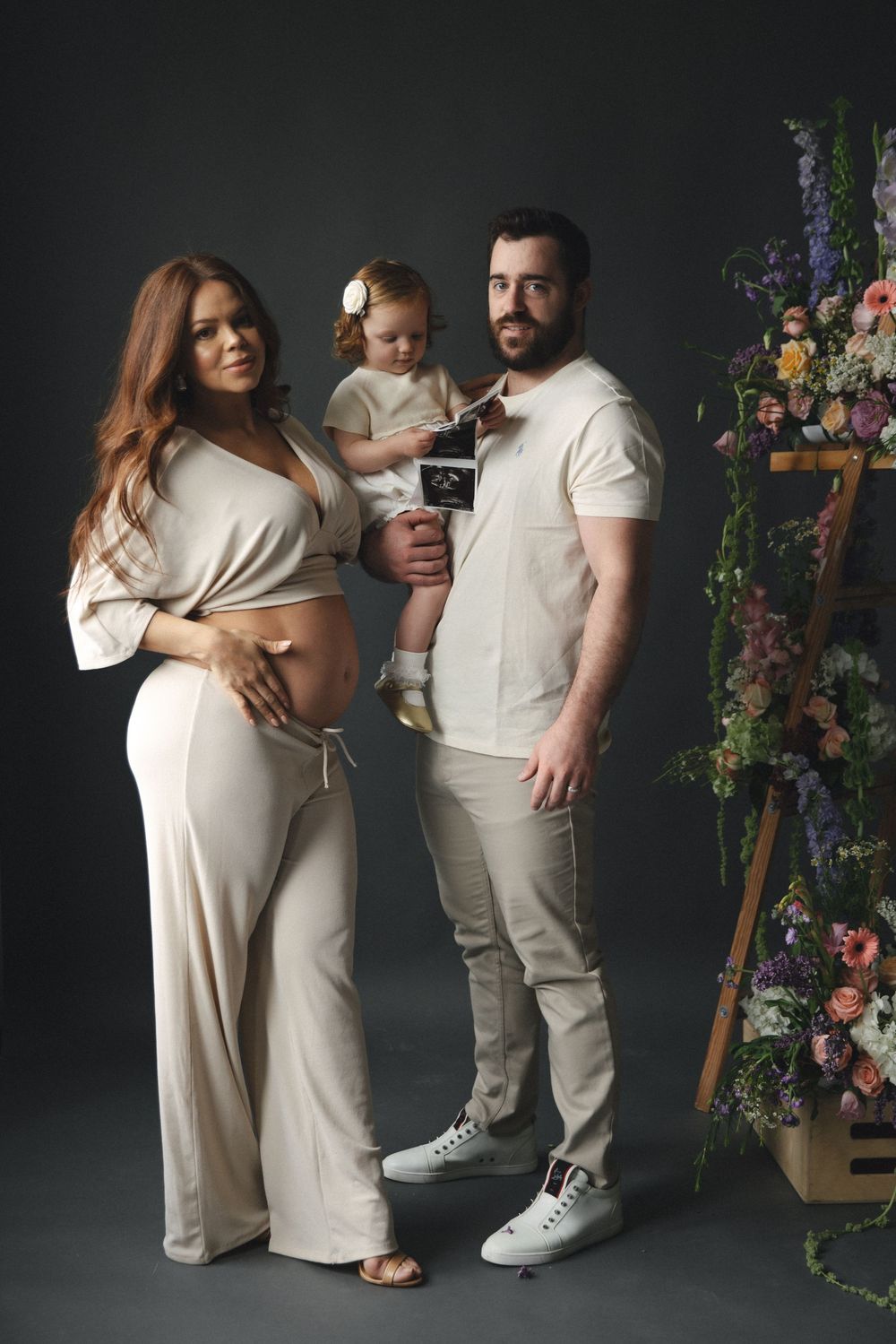 Pregnant woman in beige outfit with husband holding toddler displaying ultrasound photo beside floral arrangement.