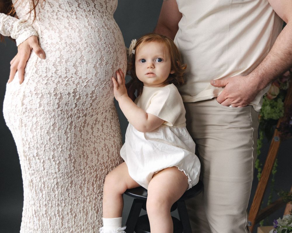 A toddler in white romper sits between pregnant mother in lace dress and father during maternity photo session.