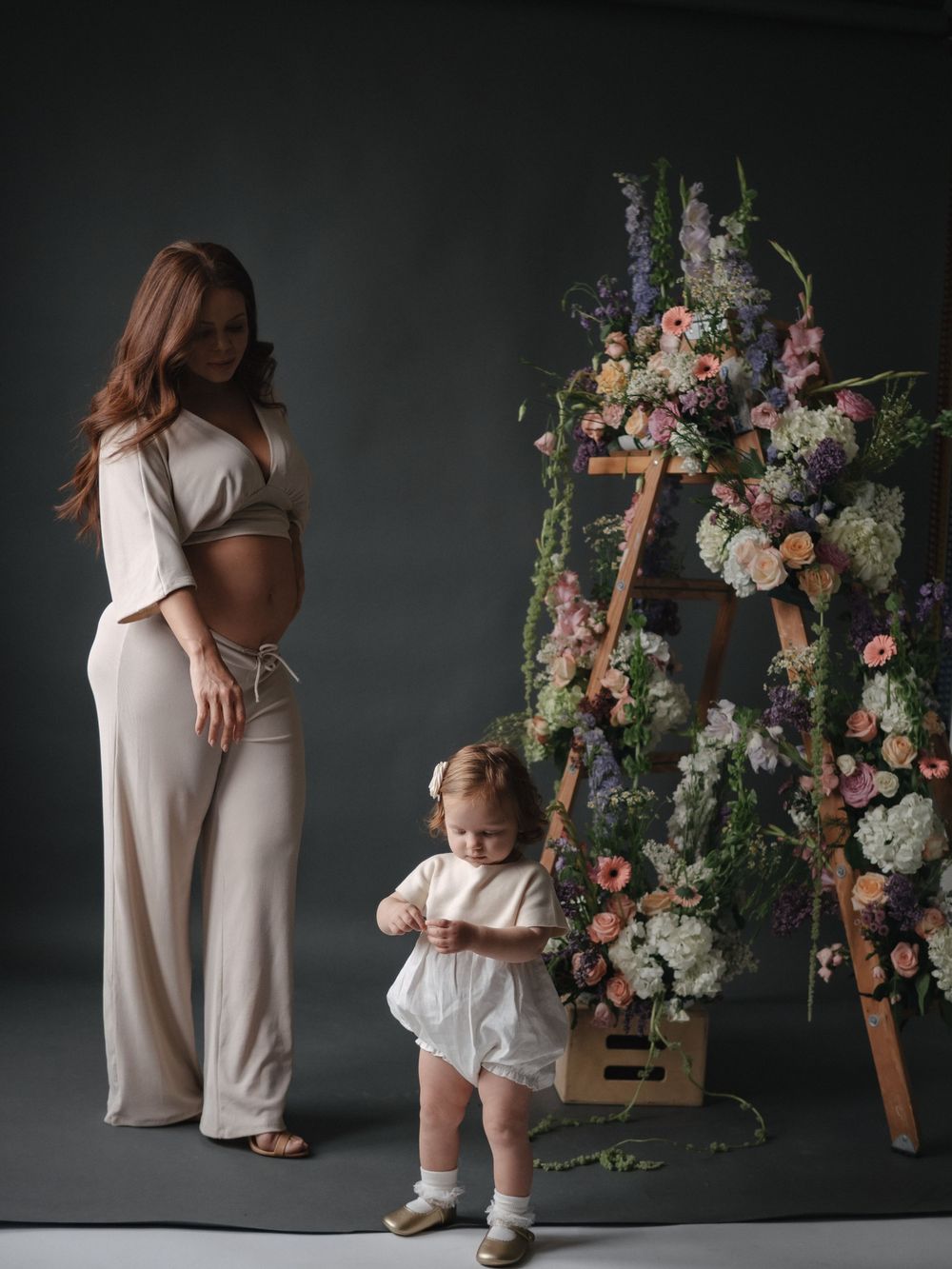 Pregnant woman in beige outfit poses with toddler girl beside a floral ladder display in a studio.