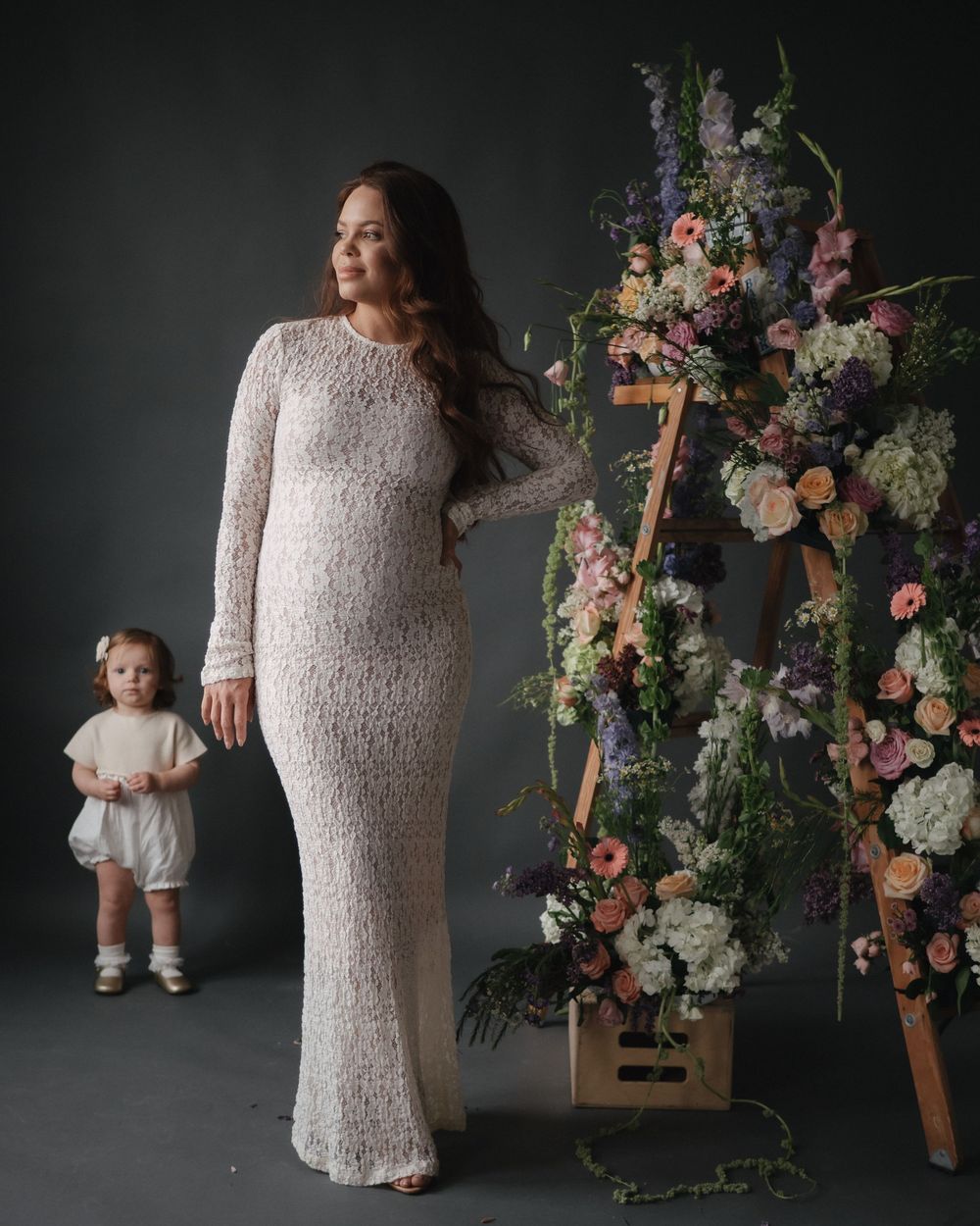 Pregnant woman in white lace gown stands beside toddler girl and floral arrangement on dark background.