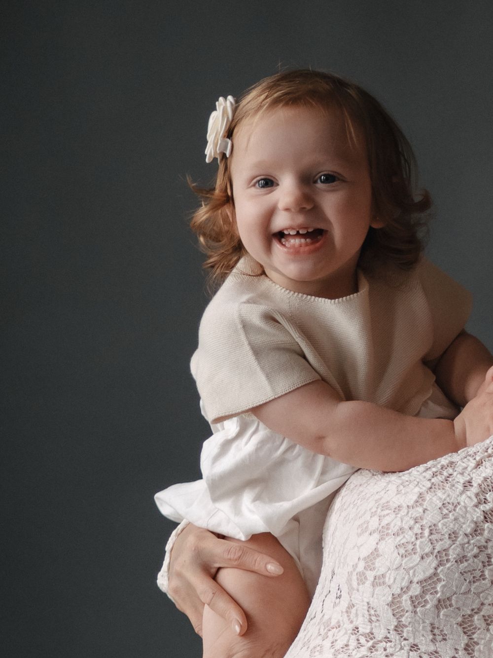 Smiling toddler girl with red curly hair and white flower clip, wearing cream dress, held by adult in lace outfit.