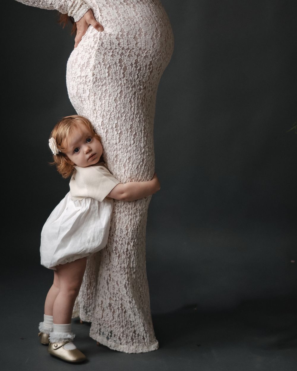 A toddler girl in a white dress hugs a pregnant woman's belly during a maternity photo session.