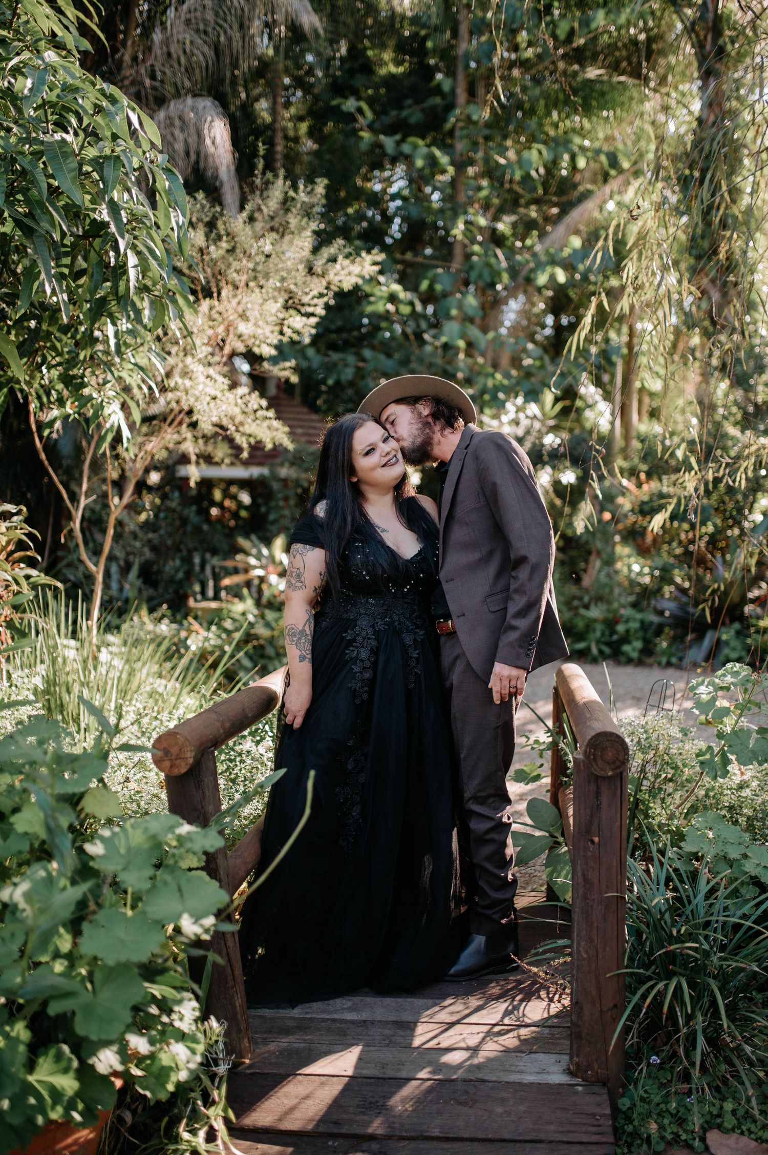 Couple in black gown and brown suit share a romantic kiss on a wooden bridge surrounded by lush greenery.