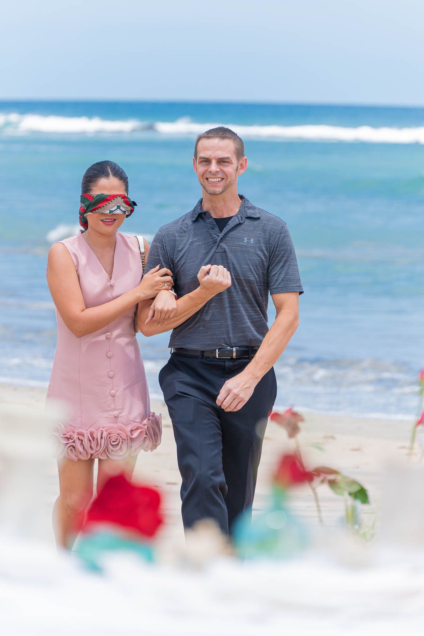 Couple walking hand-in-hand on a beach with red roses and candles in the foreground near the ocean.