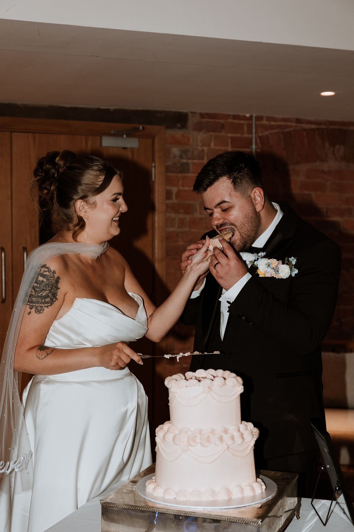 Bride and groom cutting white two-tier wedding cake, sharing a bite together in a warmly lit reception venue.