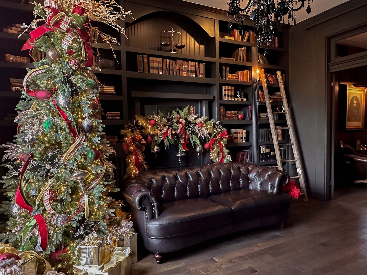 Elegant library with dark bookshelves, leather Chesterfield sofa, rolling ladder, and decorated Christmas tree with red ribbons.