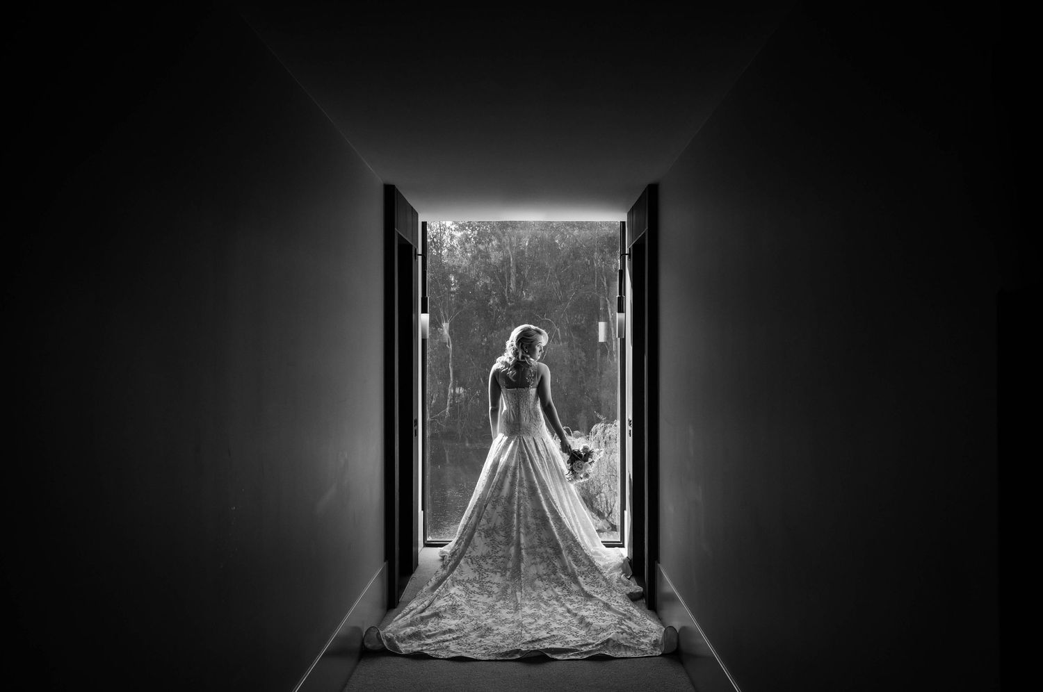 Black and white photo of a bride in a flowing gown standing at the end of a dramatic dark hallway.