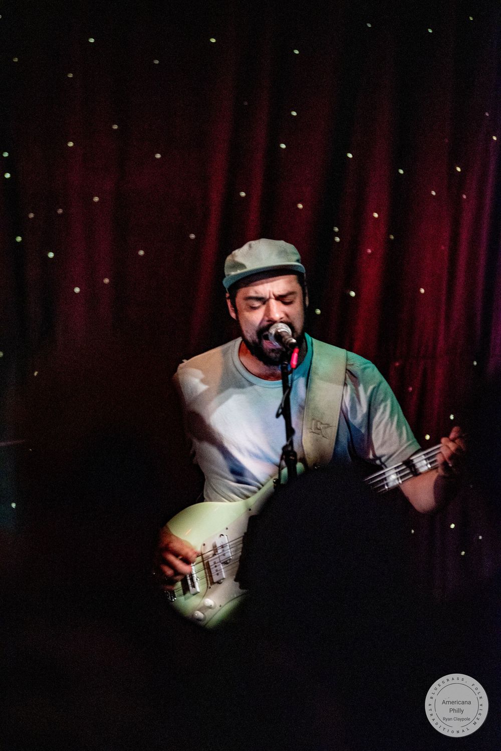 Bearded male musician wearing a cap performs on stage singing into a microphone while playing bass guitar under red curtain lighting.
