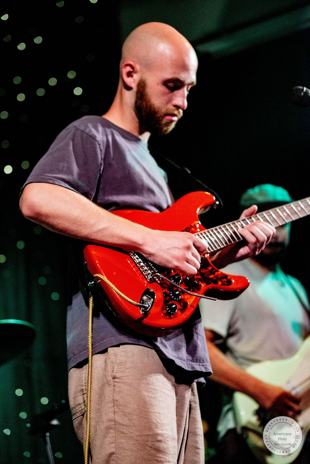 Bald guitarist with beard playing red electric guitar on stage wearing gray t-shirt and khaki pants.