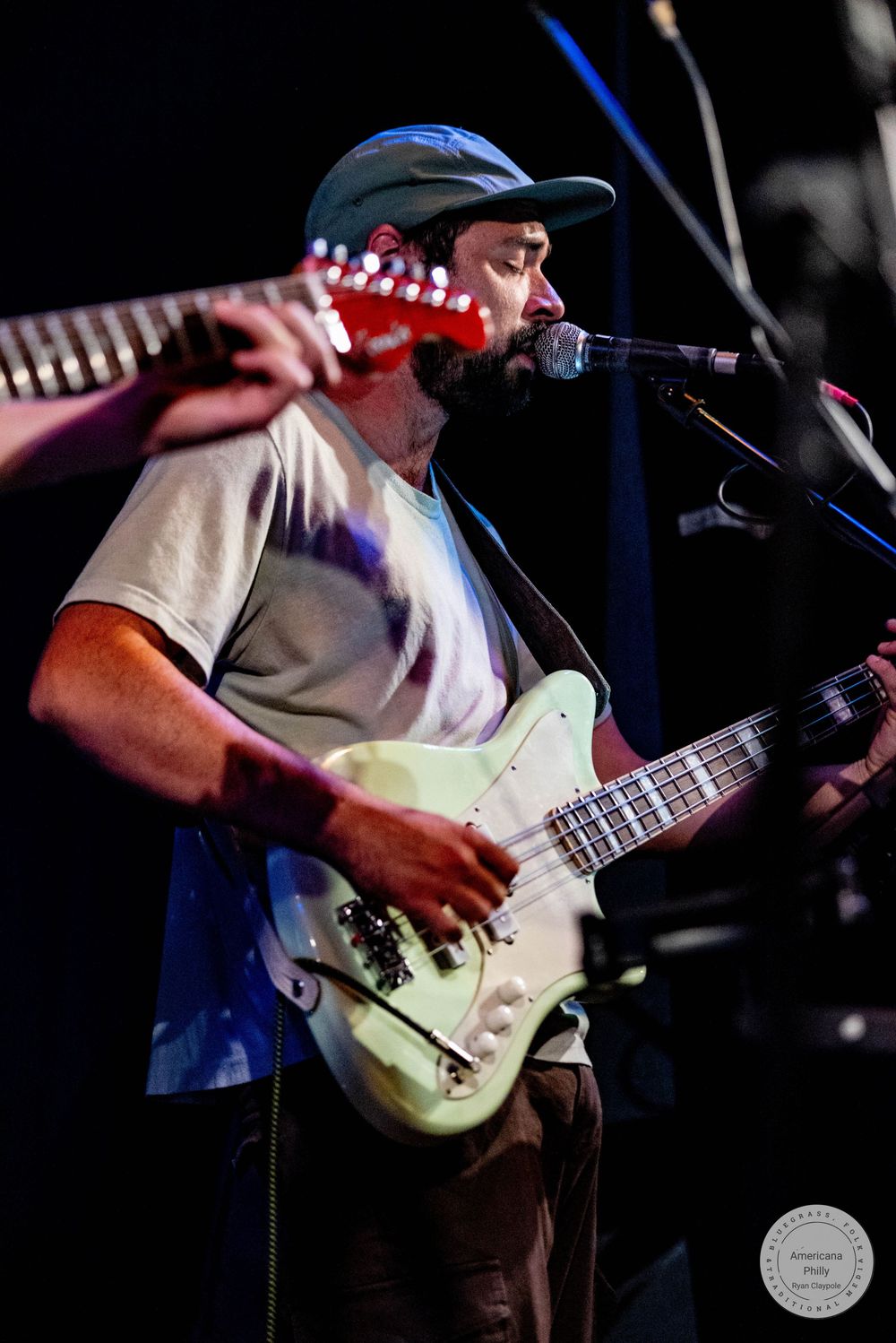 Bearded male guitarist singing into microphone while playing white electric guitar on stage at live music concert.