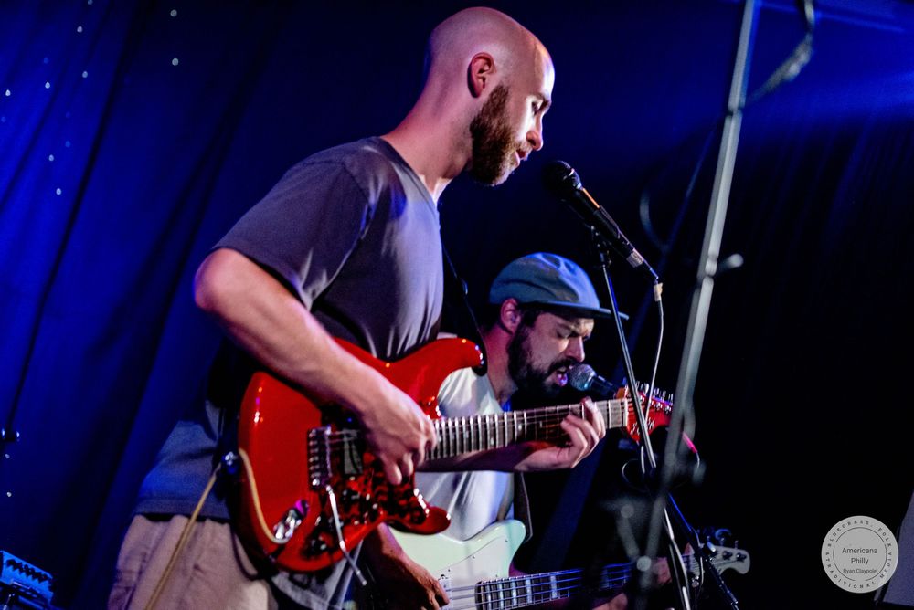 Two musicians performing live on stage, one playing a red electric guitar at a microphone under blue stage lighting.