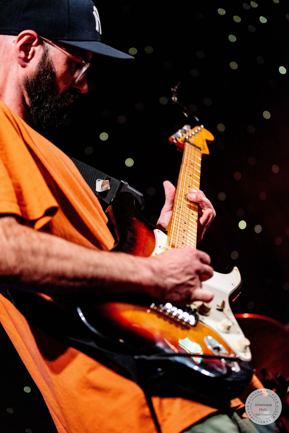 Bearded musician in orange shirt and black cap plays sunburst electric guitar on stage under bokeh lights.