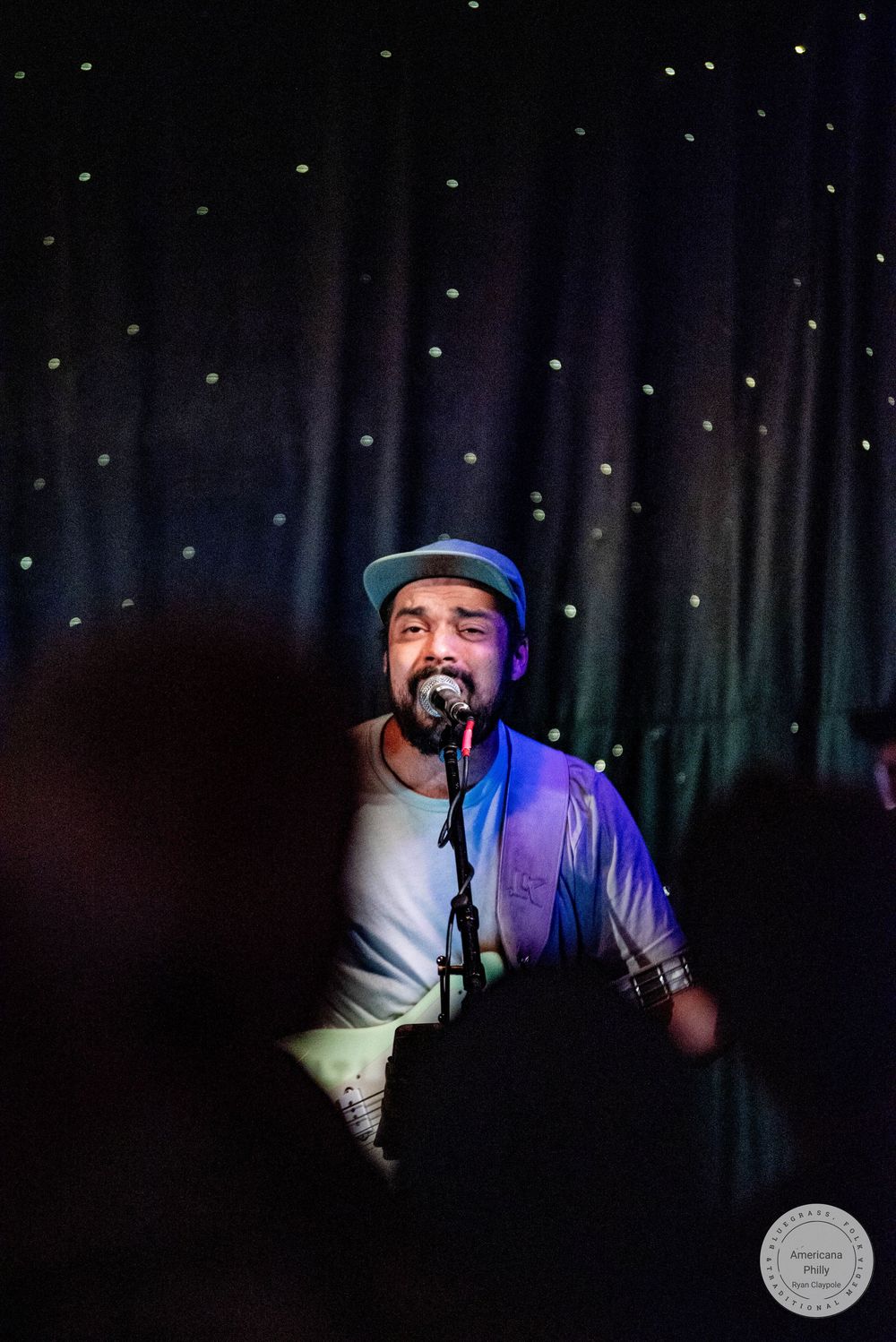 A male singer performs on stage wearing a cap and white shirt, singing into a microphone against a dark starry backdrop.