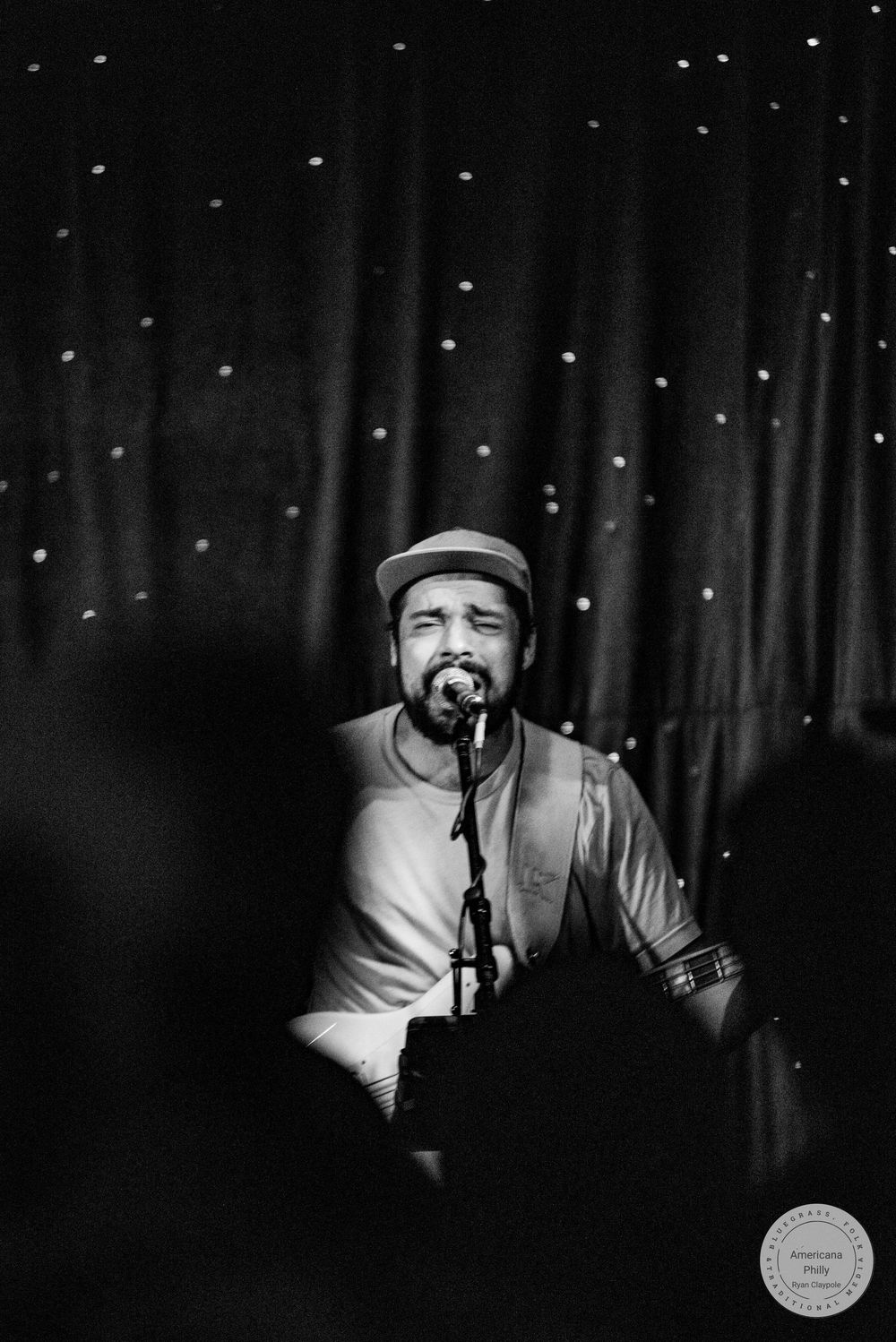 Black and white photo of a male singer performing on stage with a microphone, wearing a cap under a starry curtain backdrop.