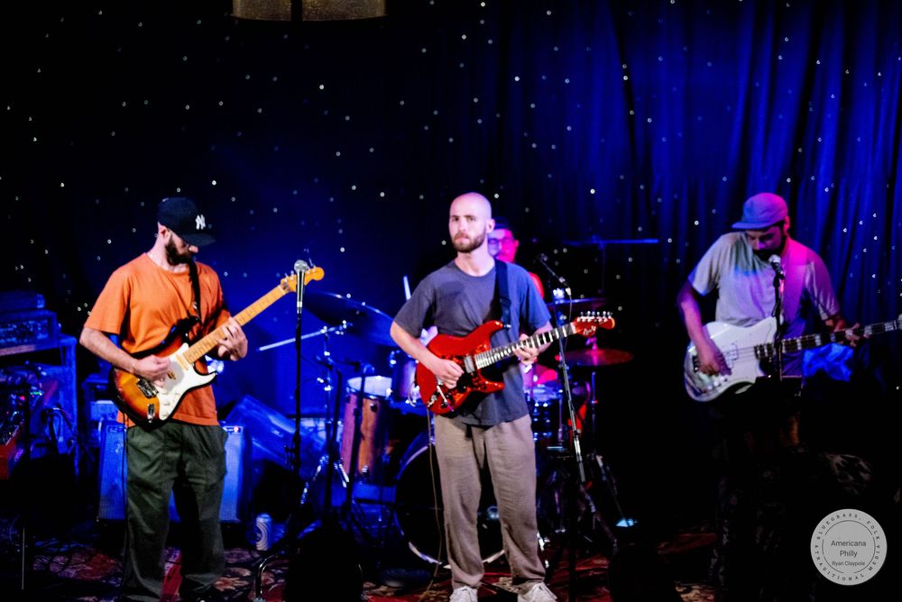 Three musicians performing on stage with electric guitars under blue starry backdrop lighting at a live concert.