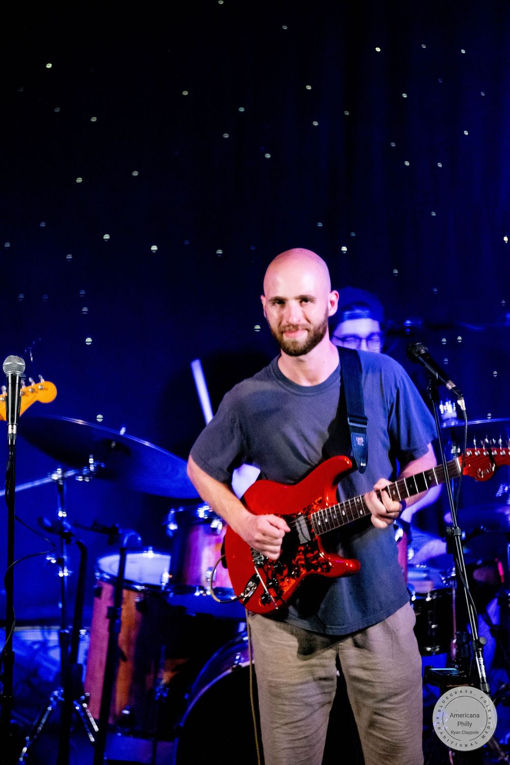 Bald guitarist with beard playing red electric guitar on stage during live concert performance.