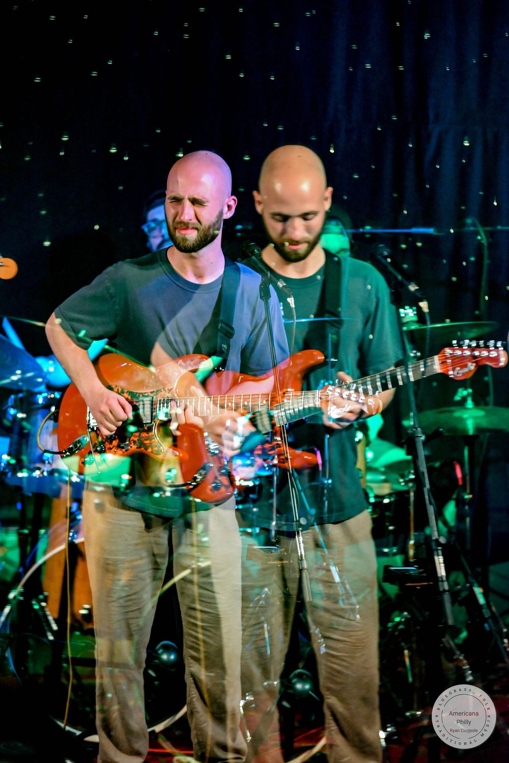 Two bald bearded guitarists playing red electric guitars together on stage at live music event.