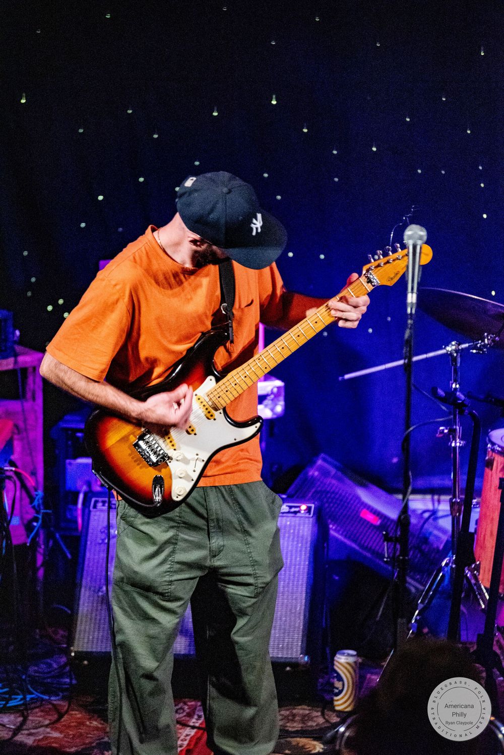 Musician in orange shirt and black cap playing sunburst Stratocaster guitar on stage with blue starry backdrop.