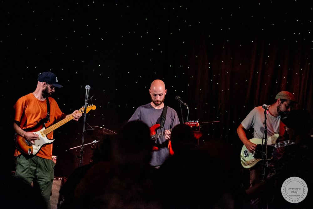 Three musicians performing on stage with guitars under starry backdrop lights in a dimly lit venue.
