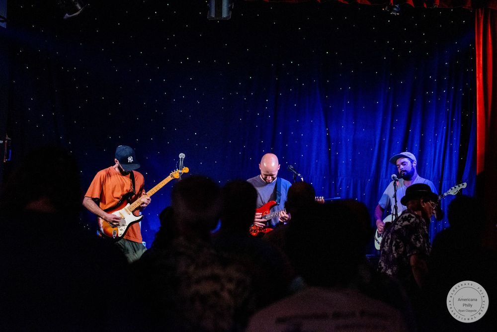 Three musicians performing on a dimly lit stage with blue star backdrop at a live concert venue.