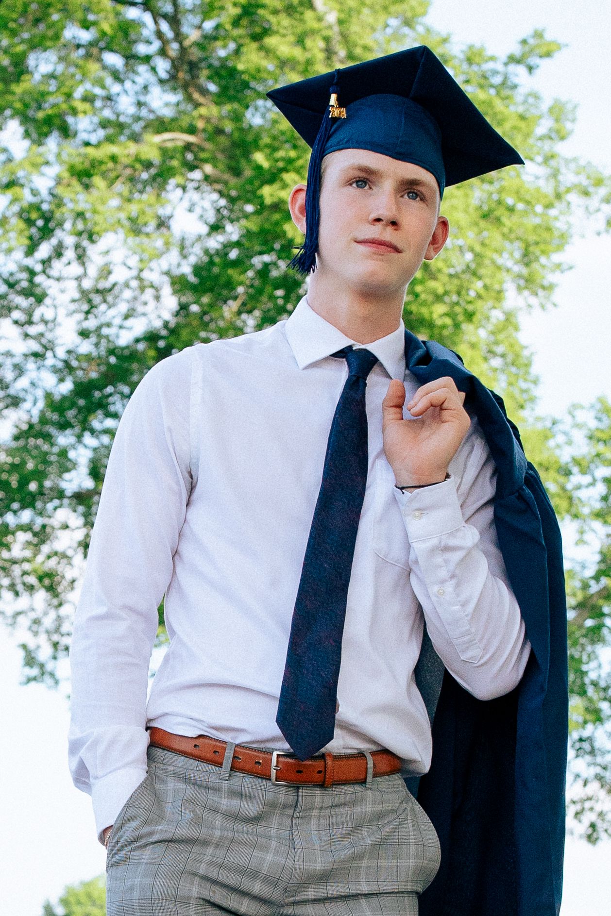 Young man in graduation cap and navy tie holds gown over shoulder outdoors, celebrating academic achievement.