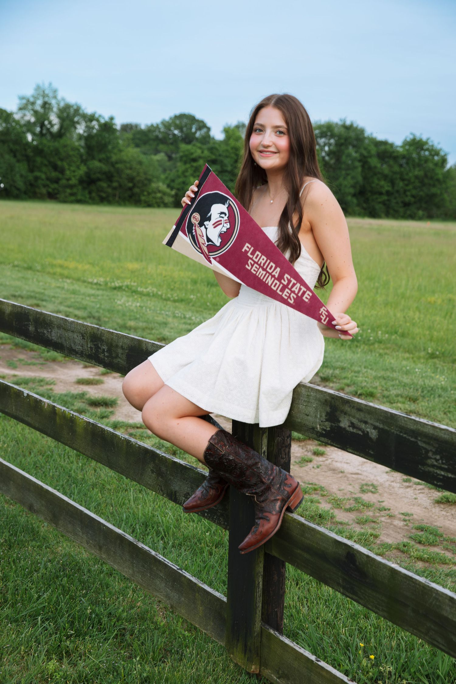 Young woman in white dress and cowboy boots holding a Florida State Seminoles pennant while sitting on a wooden fence.