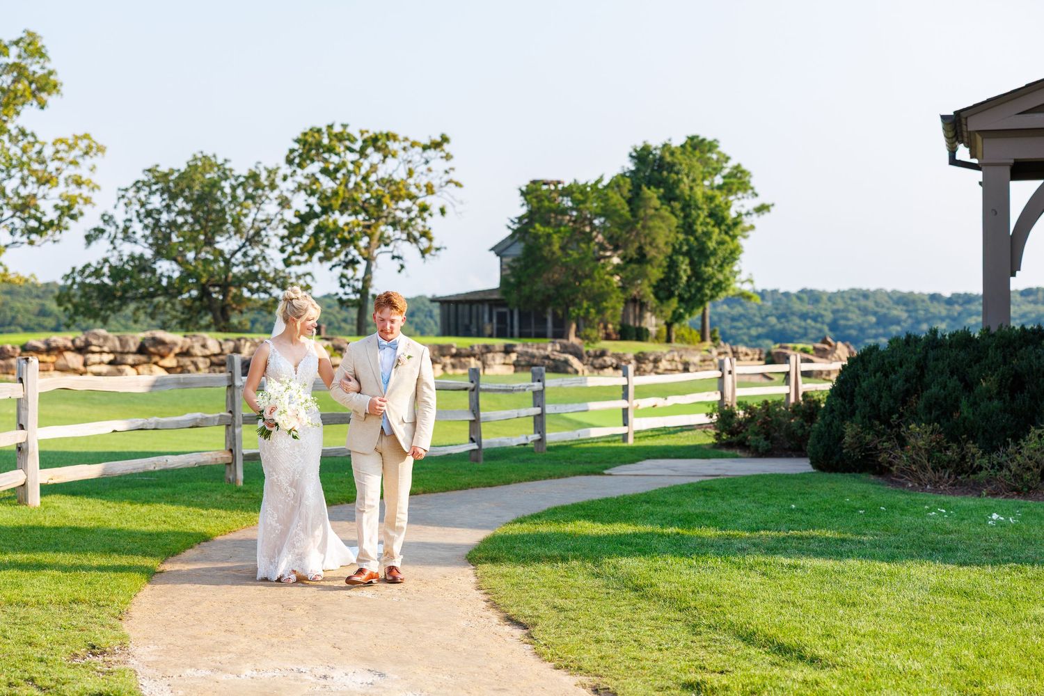 Bride and groom walking hand in hand on a garden path at an outdoor wedding venue with white fences and green trees.