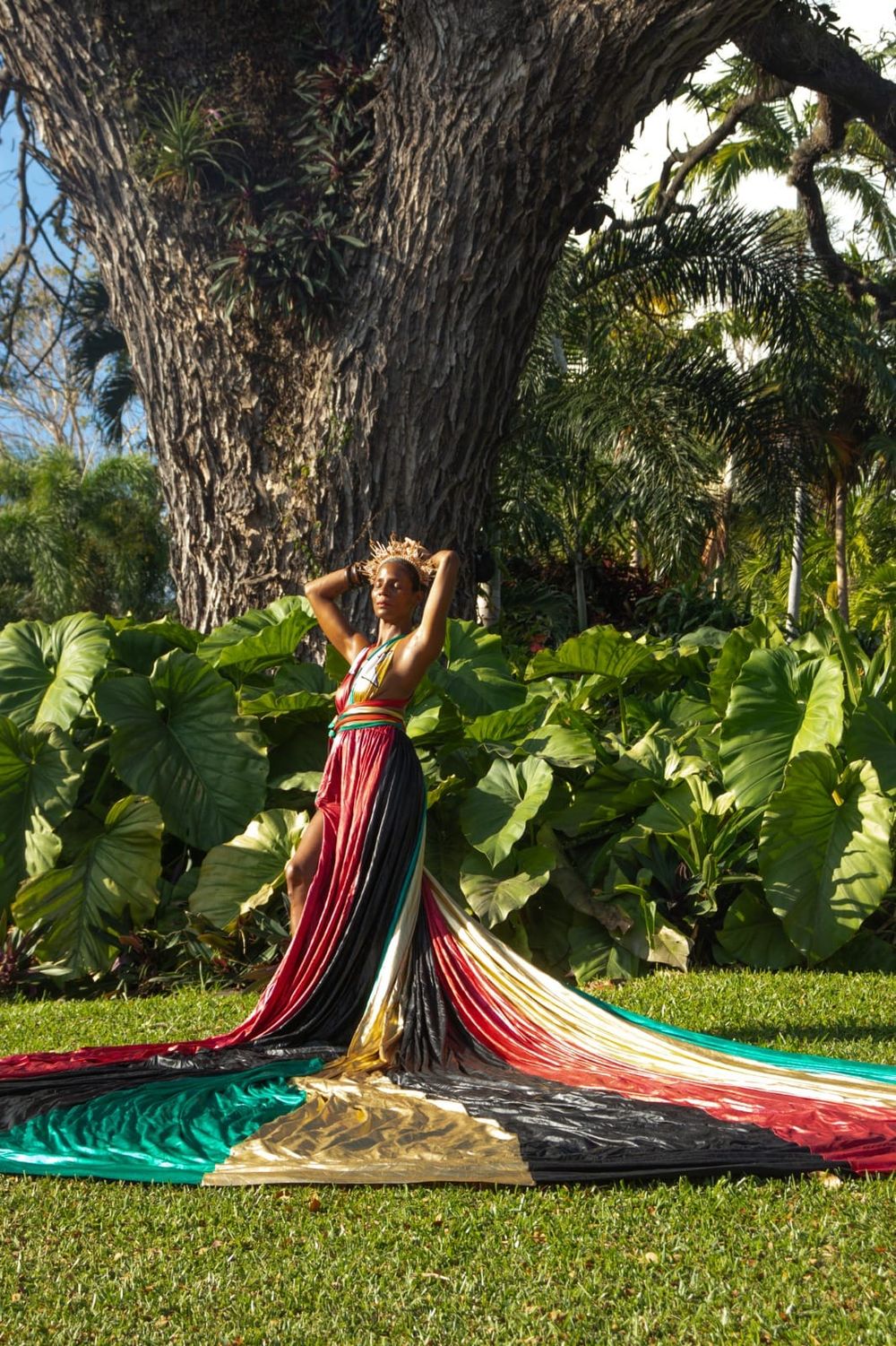 Woman in colorful flowing gown poses gracefully before a large tropical tree surrounded by lush green foliage.