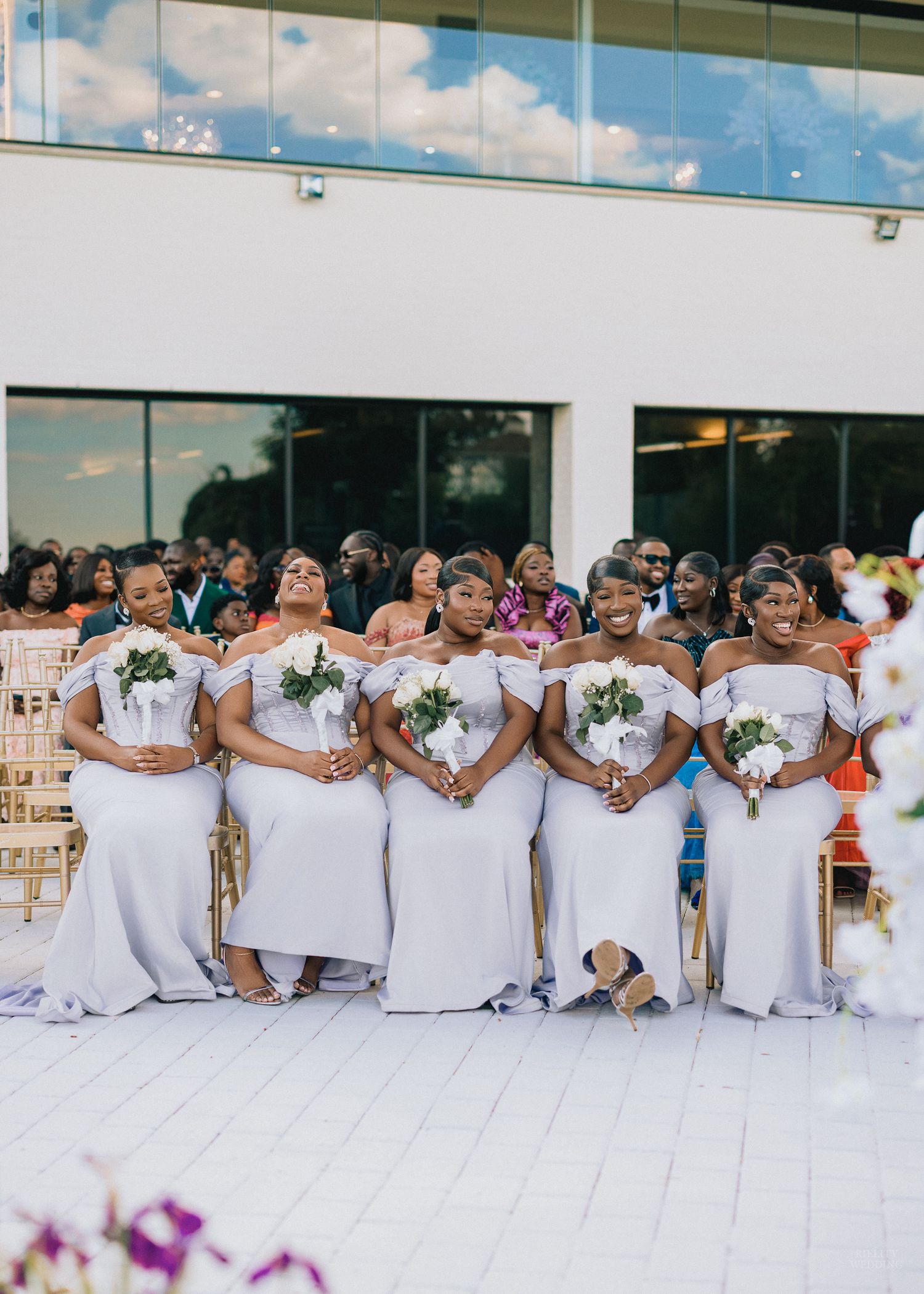 Five bridesmaids in matching white off-shoulder gowns holding bouquets at an outdoor wedding ceremony.
