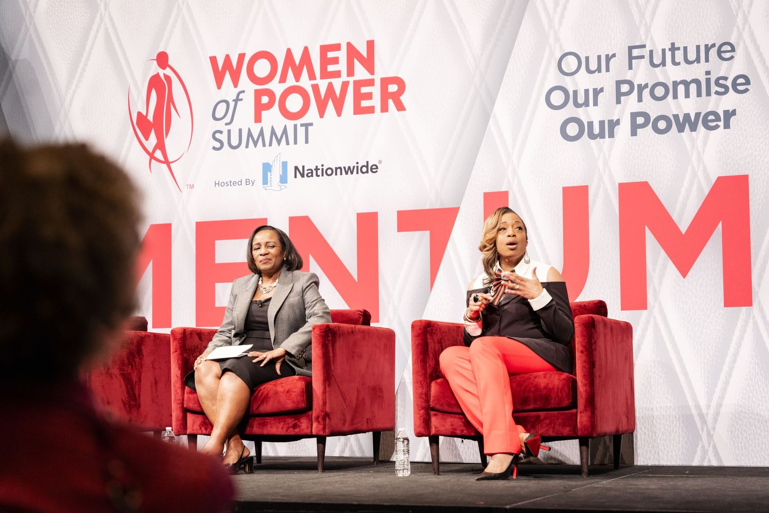 Two women speak on stage at the Women of Power Summit, sponsored by Nationwide, seated in red chairs.