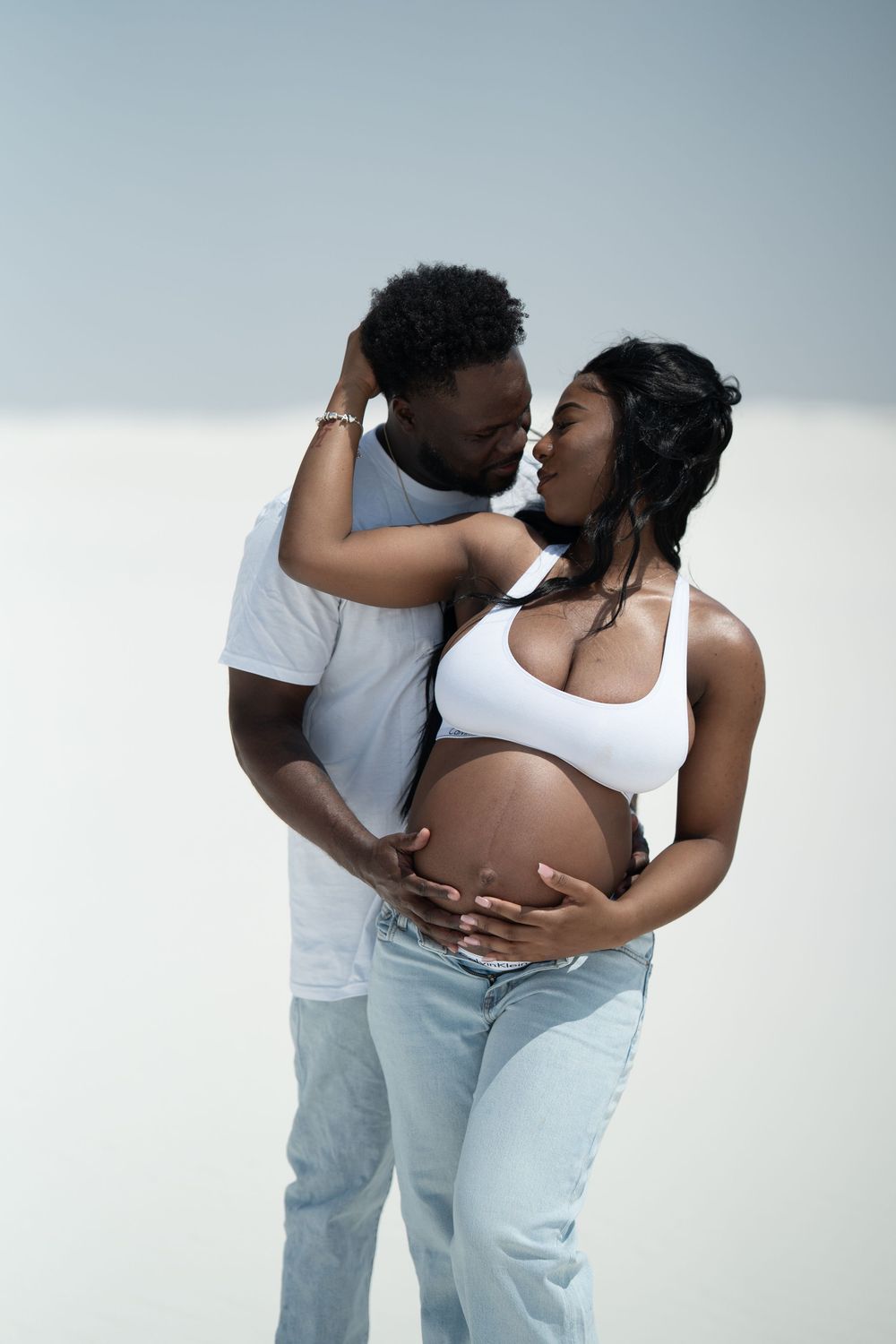 A pregnant Black couple shares a tender moment, the man embracing the woman from behind while cradling her baby bump.