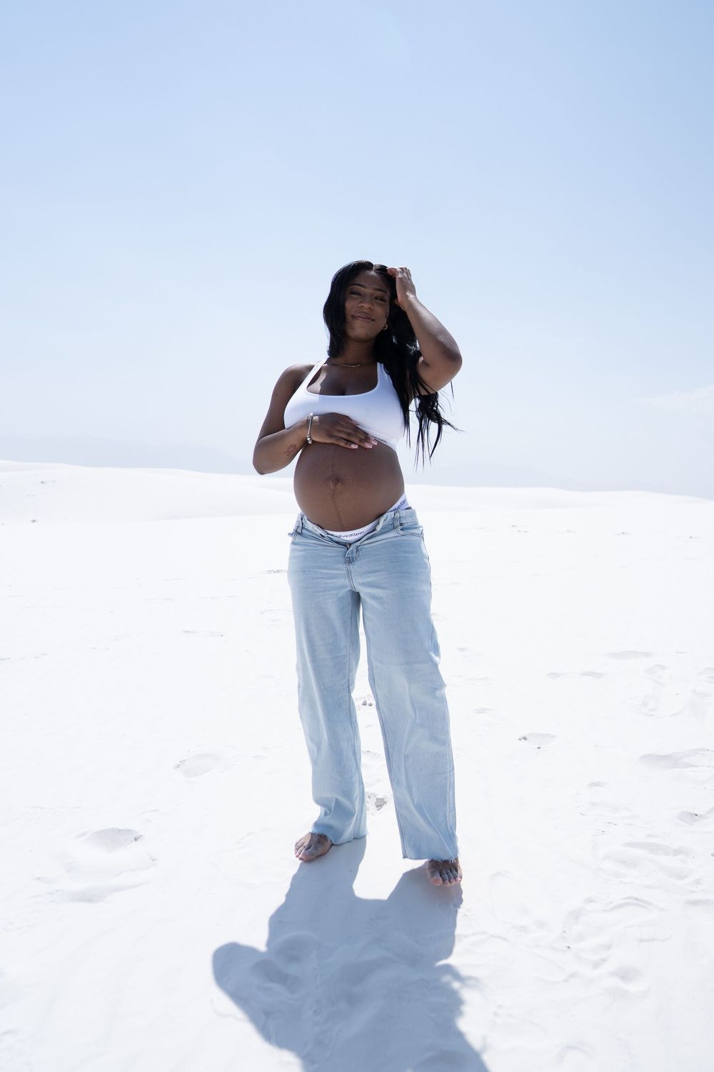 Pregnant Black woman in white tank top and jeans cradles baby bump on white sand dunes.