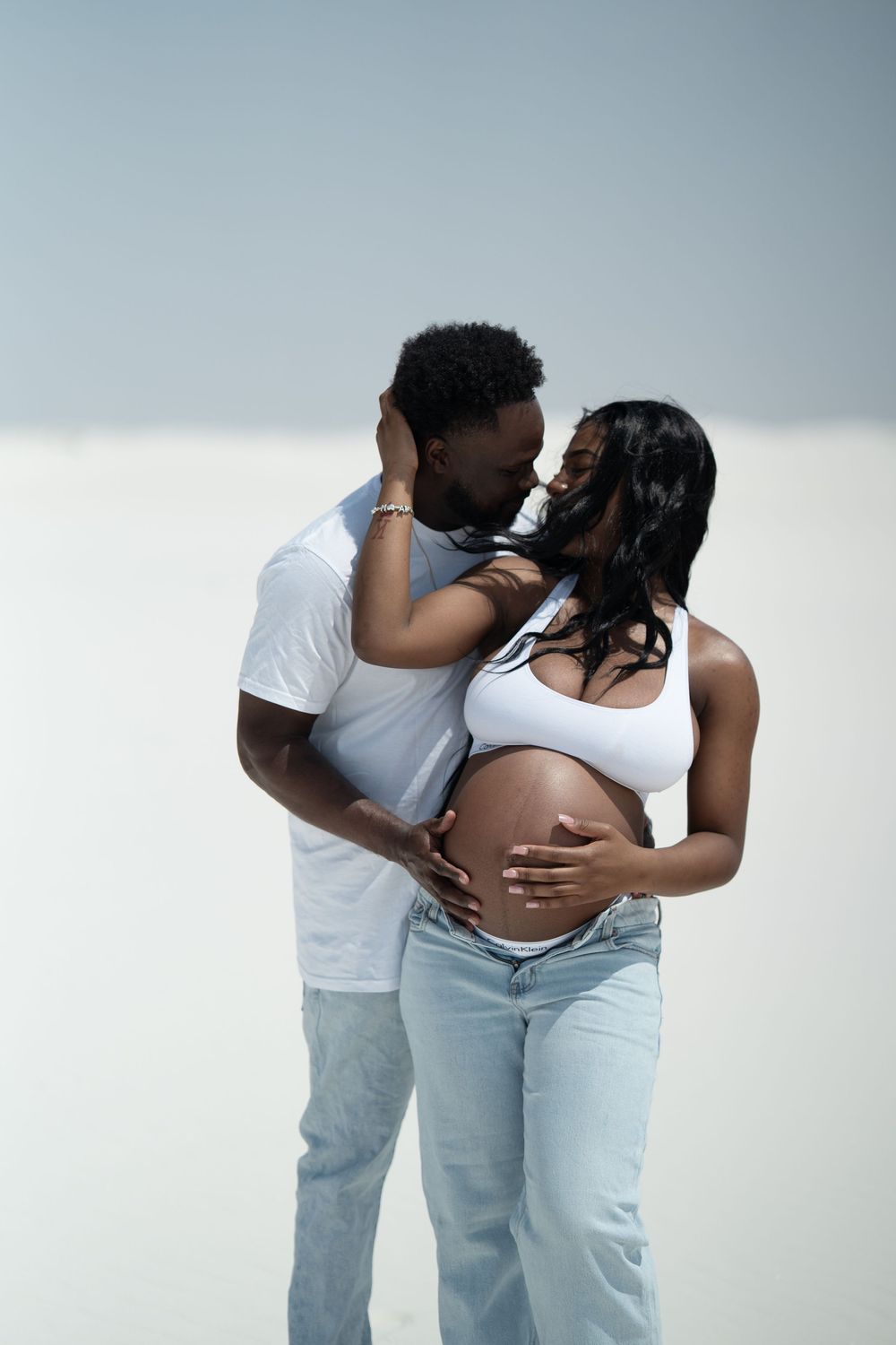 A pregnant Black woman and her partner share a tender kiss while cradling her baby bump in a bright white setting.