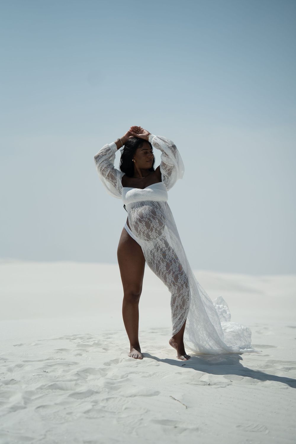 Pregnant Black woman posing in flowing white lace robe on bright white sand dunes under clear sky.