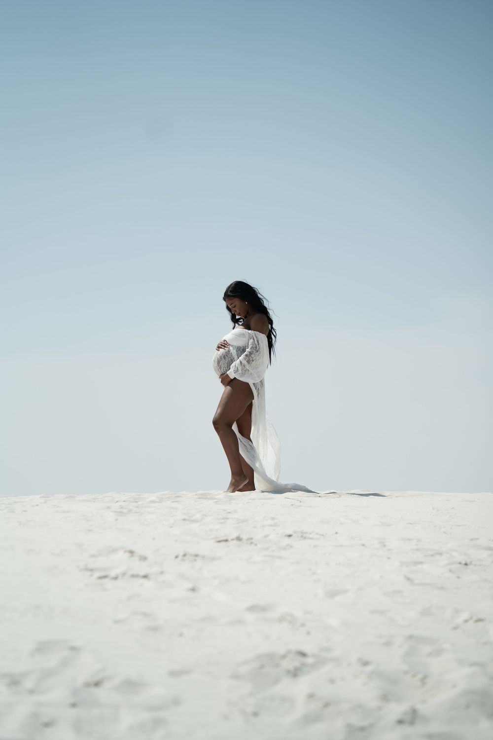 Pregnant woman in flowing white lace robe stands elegantly on white sand dunes under blue sky.