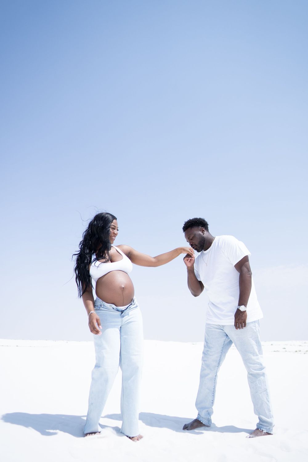 A Black man kisses a pregnant woman's hand as they stand on white sand dunes under blue sky.