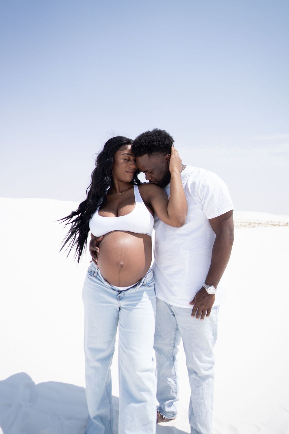 A pregnant Black woman and her partner share a tender moment on a white sand beach, both wearing white tops and jeans.