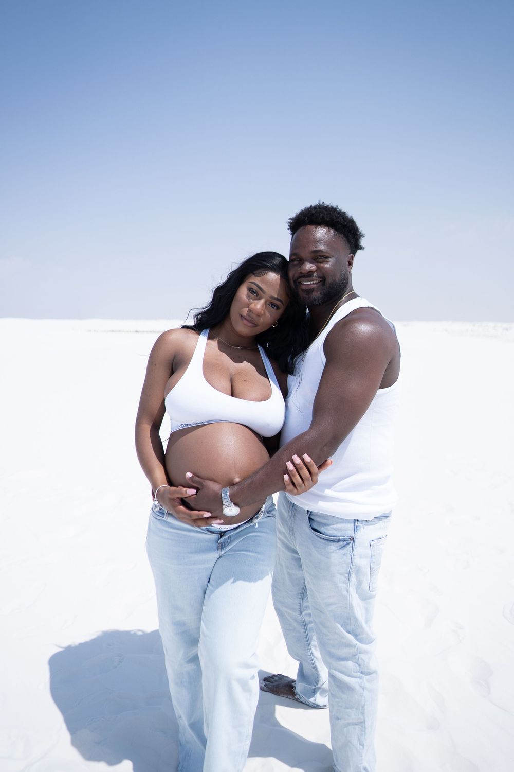 A smiling pregnant Black woman and her partner pose together outdoors, both cradling her bare baby bump on white sand.