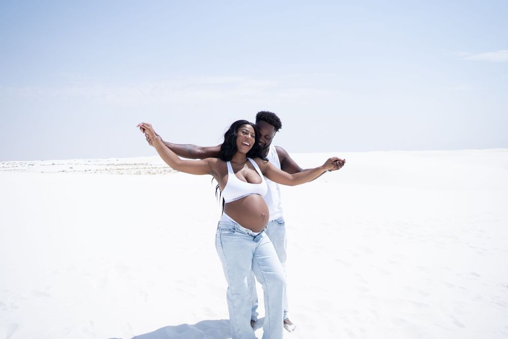 Pregnant Black couple joyfully posing in a white sand desert, woman spreading arms wide with partner behind her.
