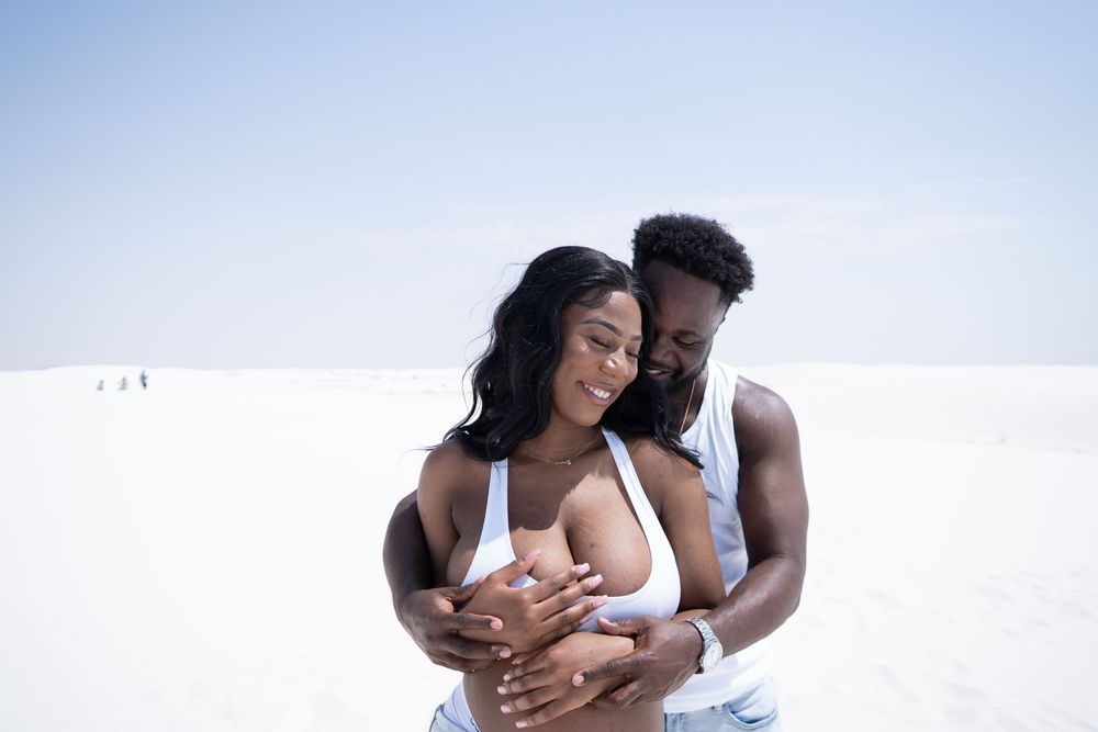 A happy Black couple embraces on a white sandy beach, smiling warmly under a clear sky.