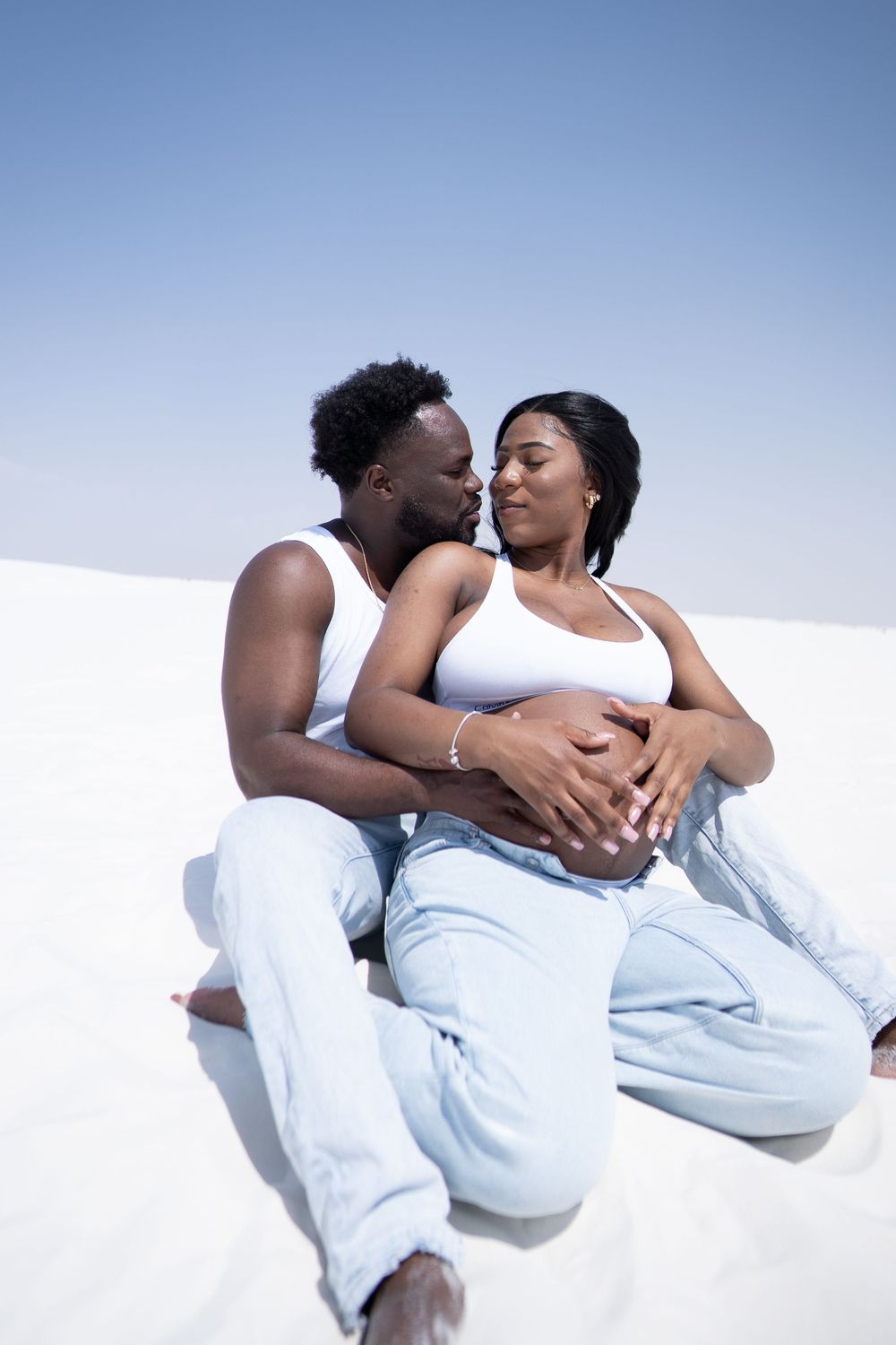 A Black couple sits together on white sand, the man kissing his pregnant partner while holding her belly.