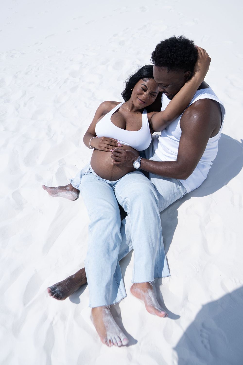 A couple sits on white sand as a man tenderly embraces his pregnant partner in a loving maternity photo.