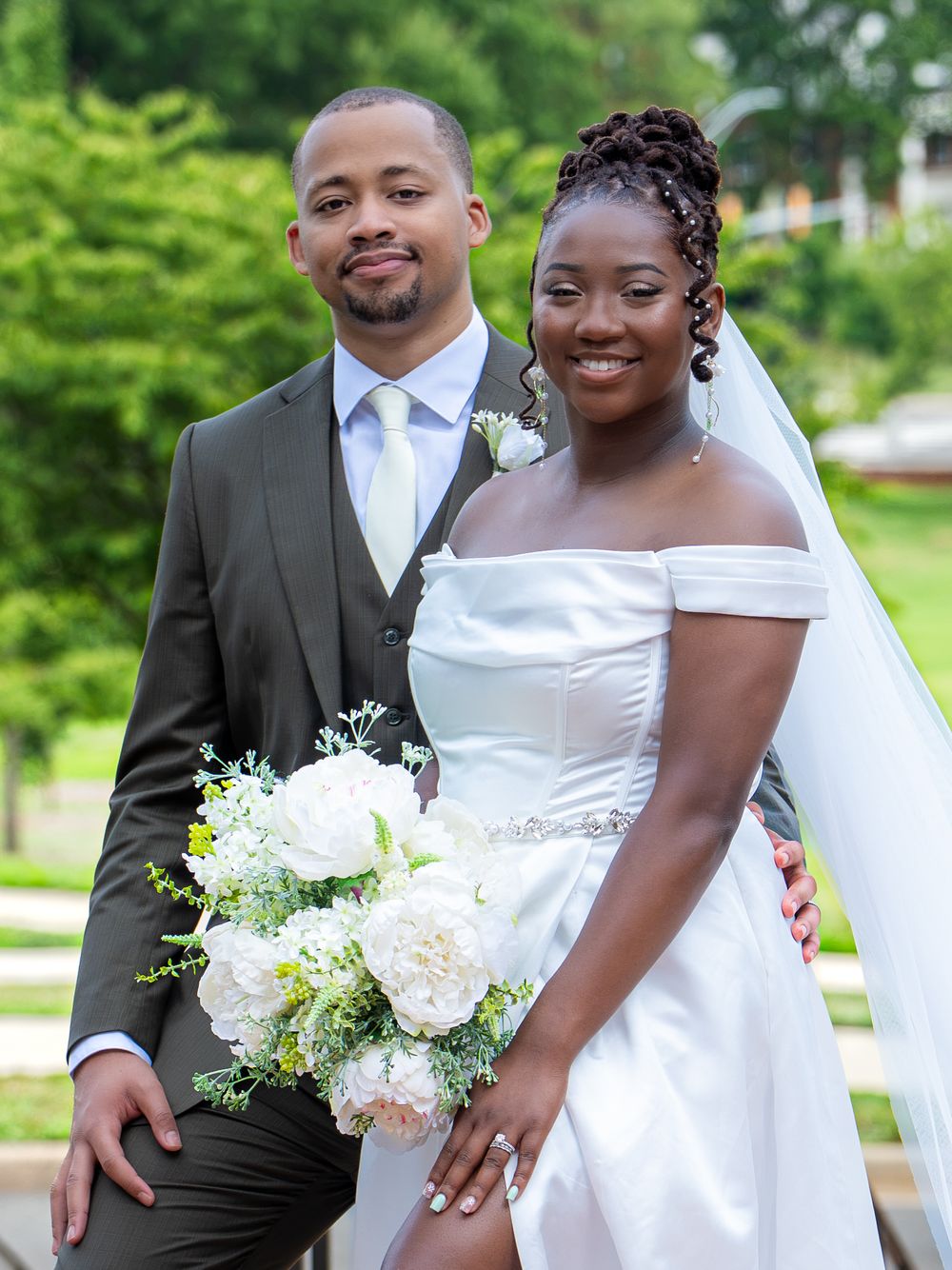 A smiling bride and groom pose together outdoors, with the bride holding a white floral bouquet on their wedding day.