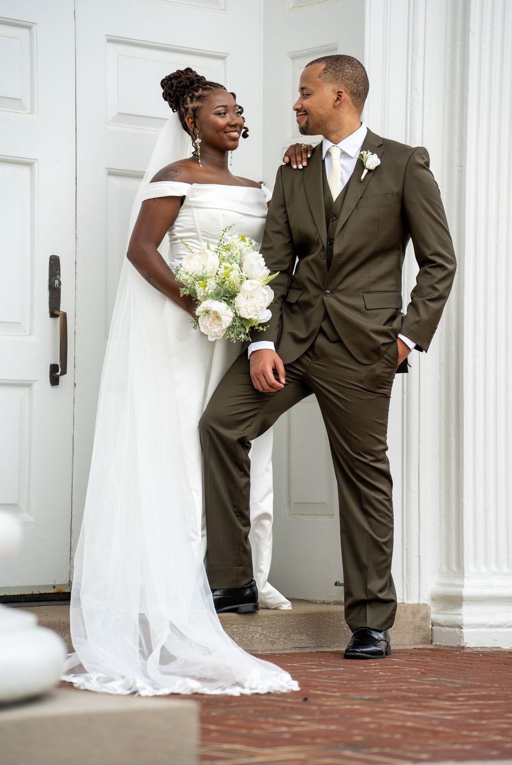 Bride in white off-shoulder gown and groom in olive suit posing at white door.