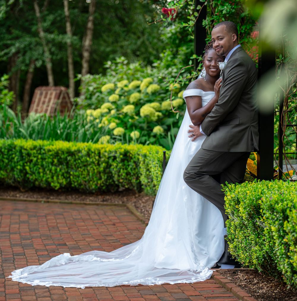 A bride and groom pose together in a lush garden, with the bride's long white train spread across brick pavers.